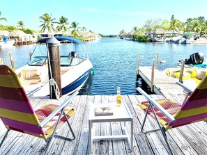 Una terraza de madera con dos sillas a rayas, una mesa con una botella y un libro, y un barco amarrado en un puerto deportivo con palmeras y chozas de techo de paja al fondo.