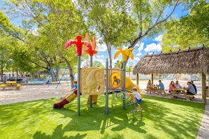 Ein Spielplatz mit Rutschen und Klettergeräten, Picknicktischen und einem strohgedeckten Unterstand.