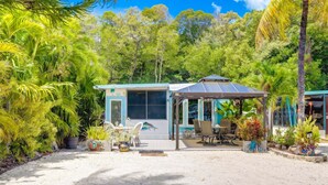 Una pequeña casa moderna con un porche cubierto, rodeada de exuberante vegetación tropical.