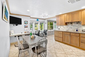 A kitchen with wooden cabinets, a tiled floor, a dining table with chairs, and a television mounted on the wall.