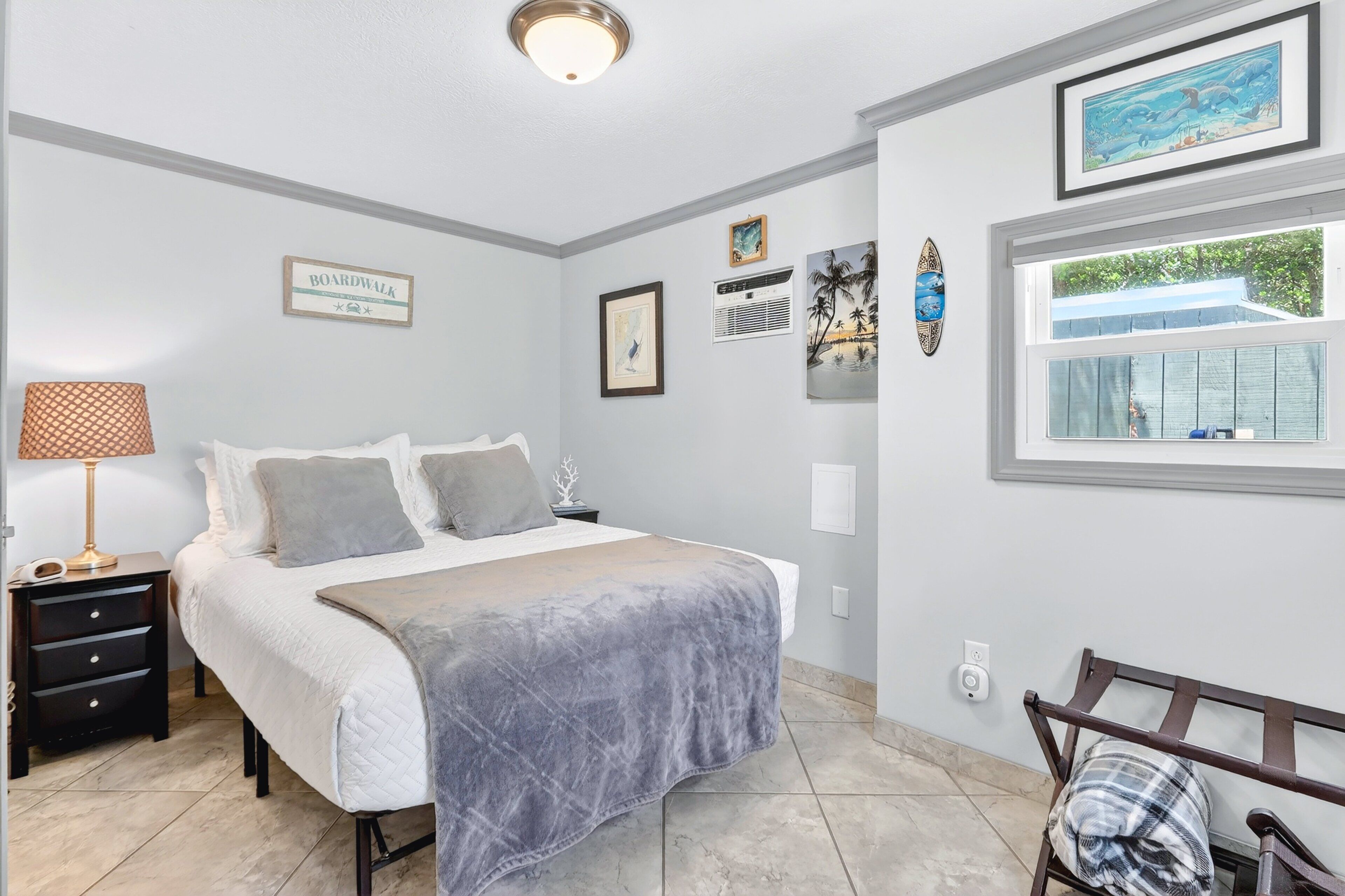 Une chambre à coucher comprenant un lit, une table de chevet, une lampe, des œuvres d’art murales et une fenêtre donnant sur un paysage.