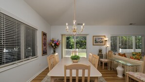 Un espace repas comprenant une longue table, des chaises, un lustre et une vue sur l’extérieur, visible à travers une fenêtre munie de stores.