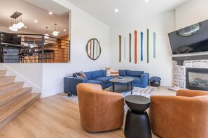 Un salon moderne avec un canapé bleu, des fauteuils orange, une cheminée et un escalier menant à un espace repas.