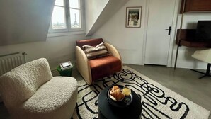 Ein gemütliches Schlafzimmer im Loft-Stil mit schrägem Dach, einem großen Fenster mit Blick auf die Stadt, einem bequemen Sessel, einem kleinen Tisch mit Frühstück und einem TV-Schrank.
