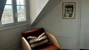 Ein Zimmer mit Blick auf eine Stadtansicht durch ein Fenster, ein gerahmtes Bild an der Wand und ein Sessel mit Kissen.
