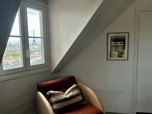 Ein Zimmer mit Blick auf eine Stadtansicht durch ein Fenster, ein gerahmtes Bild an der Wand und ein Sessel mit Kissen.