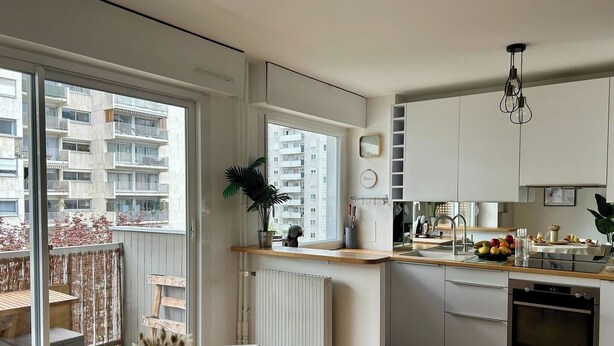 Et moderne køkken med hvide skabe, et spiseområde med et bord dækket til morgenmad, og udsigt til en boligbygning udefra.