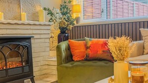 A cozy living room with a fireplace, a sofa with colorful cushions, a vase with flowers, and a window with blinds. - Forest Inspired Home! Near M1 and Superb Locations (Nottingham)