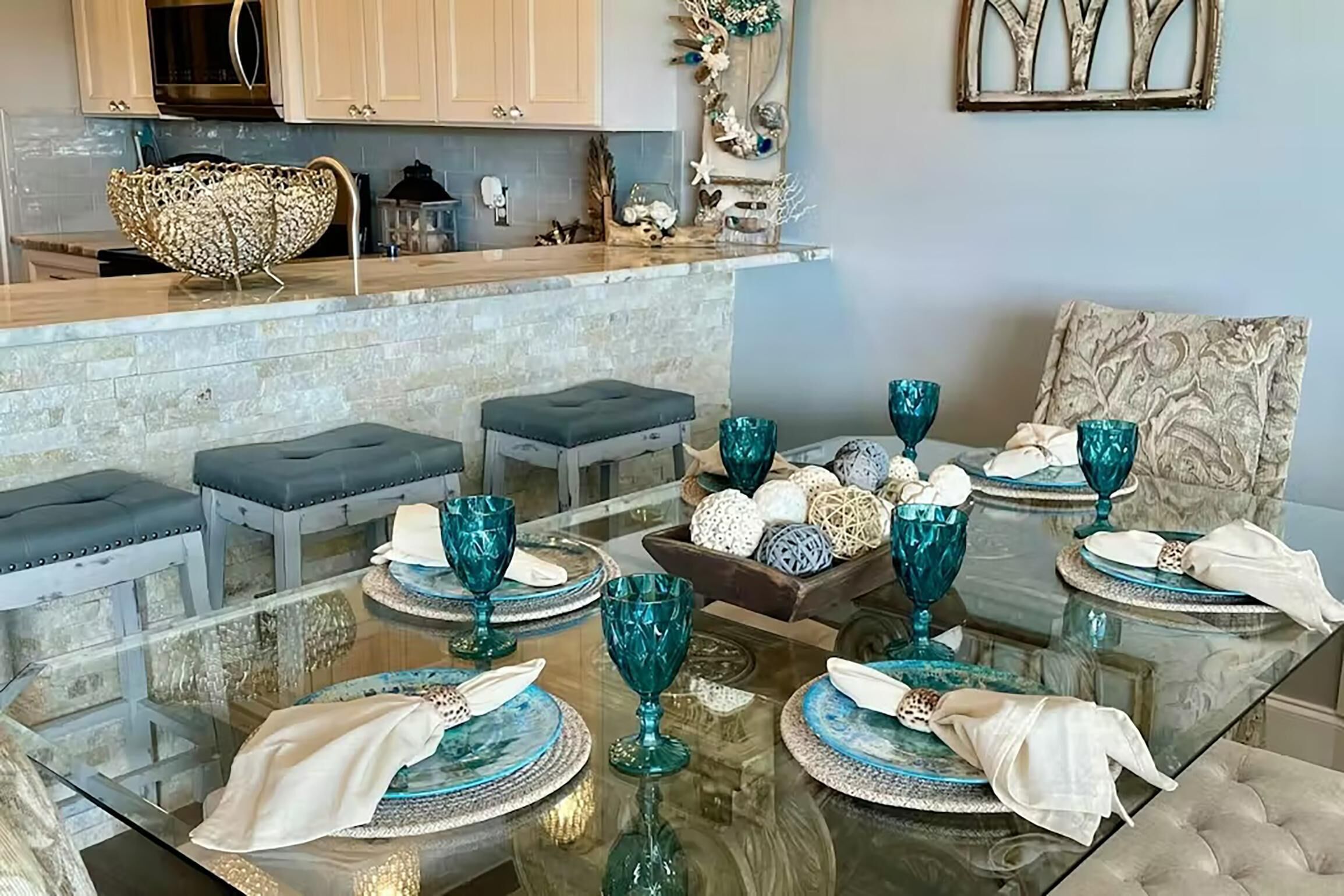 Un comedor con una mesa de vidrio dispuesta para seis personas, cristalería azul y un cuenco decorativo con huevos en la encimera.
