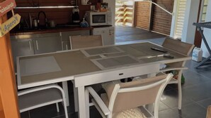 A dining area with a table, chairs, a kitchen in the background, and a wooden deck. - KaZa Bel'Vi By La Manoshra (Capesterre de Marie-Galante)