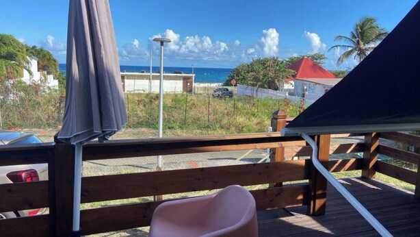 A wooden deck with a pink chair, a covered area with a table, and a view of the ocean and buildings in the background. - KaZa Bel'Vi By La Manoshra (Capesterre de Marie-Galante)