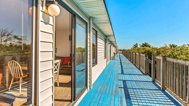 A covered outdoor deck with a blue floor, glass doors leading to a room with a chair and a table, and a wooden fence along the side. - Ocean Surf Condo | Queen Bed | Sleeps 4 | Unit 207 (Ocean Shores)