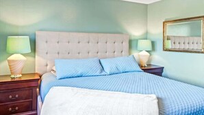 A bedroom with a tufted headboard, a bed with blue bedding, two bedside lamps, a nightstand, and a mirror on the wall. - Ocean Surf Condo | Queen Bed | Sleeps 4 | Unit 207 (Ocean Shores)