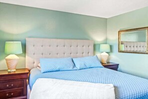 Un dormitorio con cabecera capitonada, cama con ropa de cama azul, dos lámparas de noche, una mesita de noche y un espejo en la pared.