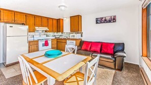 A compact kitchen with wooden cabinets, a white refrigerator, and a dining area with a table and chairs. - Ocean Surf Condo | Queen Bed | Sleeps 4 | Unit 207 (Ocean Shores)