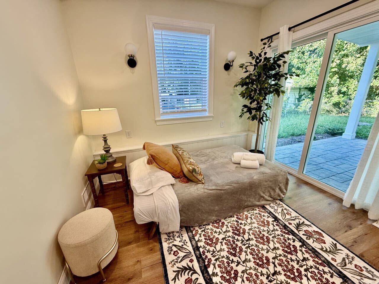 Un dormitorio con cama, mesita de noche, lámpara, silla y vistas al exterior.