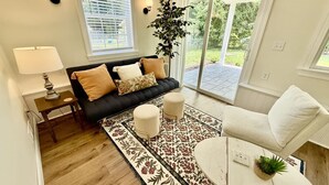 Een woonkamer met een bank, poefjes, een salontafel, een lamp, een plant en uitzicht door een glazen deur.