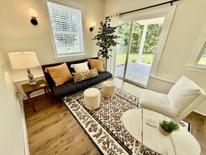 A living room with a sofa, ottomans, a coffee table, a lamp, a plant, and a view through a glass door.