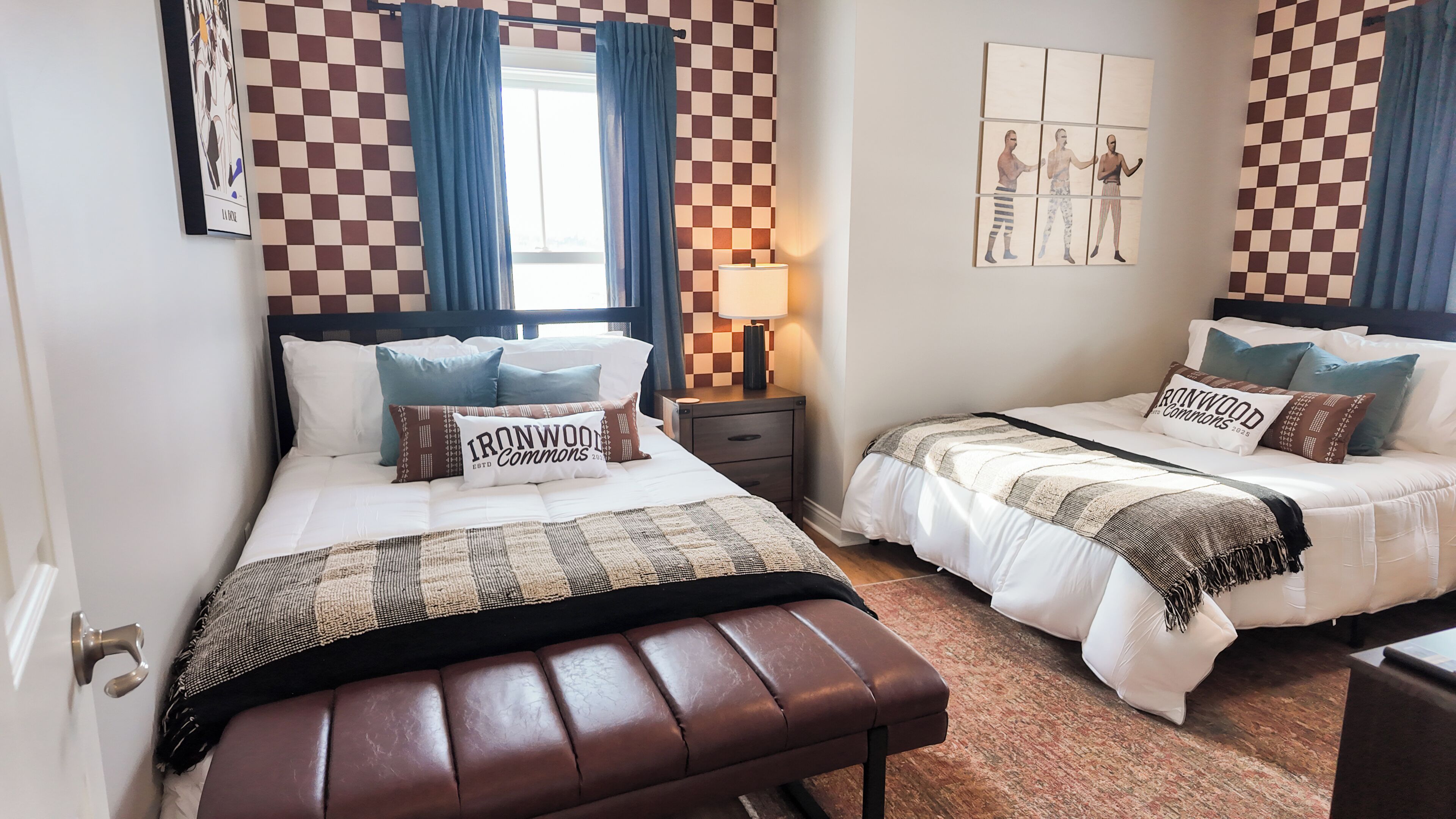 Een hotelkamer met twee bedden, wanden met een geruit patroon, een raam met blauwe gordijnen, een nachtkastje met een lamp en een kunstwerk aan de muur.