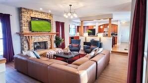 A living room with a stone fireplace, leather sofa, and a television mounted on the wall. - Sleep 30, Near Grand Park, Golf Sim, Sauna, Hottub (Westfield)