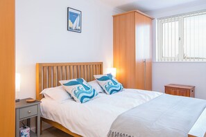 A bedroom with a wooden headboard, a bed with white bedding and blue patterned pillows, a bedside table with a lamp, a small wooden cabinet, and a framed picture on the wall. - Larus, Holiday Apartment Smoke House Quay, Milford Marina, Sleeps 4, 2 bedrooms,  bathrooms (Milford Haven)