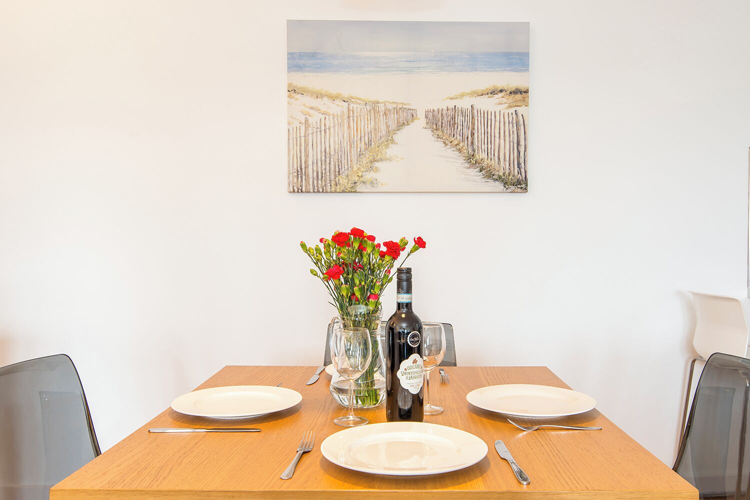 A dining table set with plates, cutlery, a vase with flowers, and a bottle of wine, with a beach path painting above.