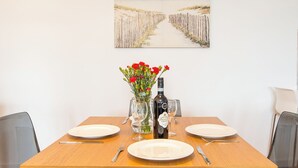Mesa de jantar posta com pratos, talheres, um vaso com flores e uma garrafa de vinho, com uma pintura de um caminho na praia acima.