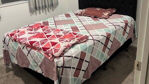 A bed with a patterned bedspread, a dark headboard, and a window with curtains. - Room-1 Spacious Bedroom Available (Papakura)
