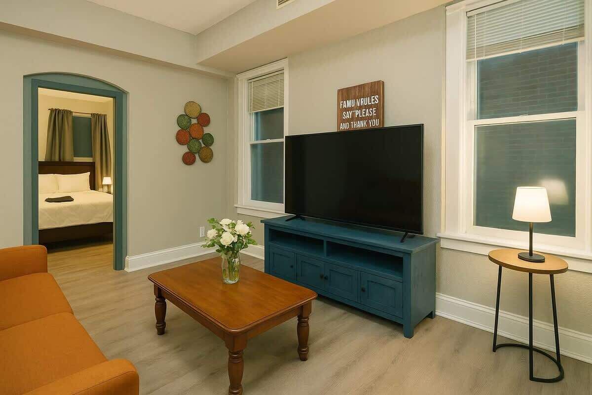 Ein Hotelzimmer mit Bett, Fernseher, Couchtisch mit Blumen, Sofa und einer Wandtafel.