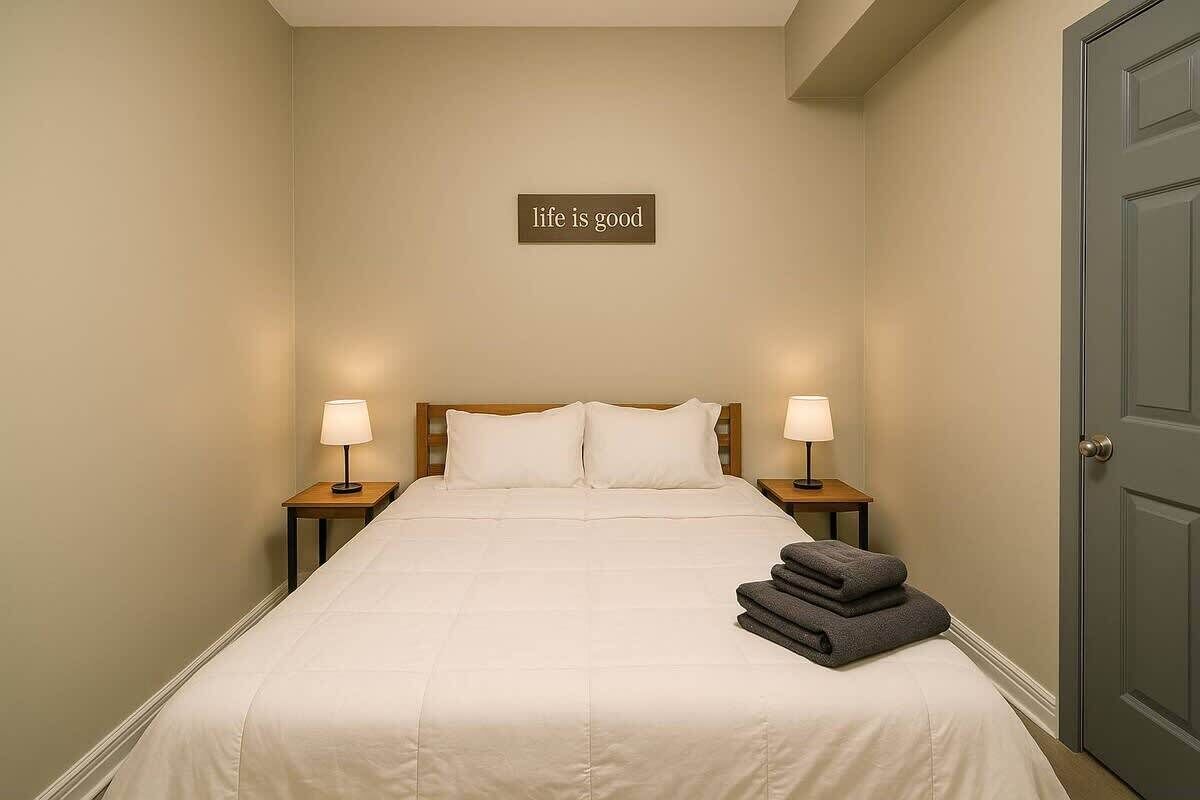 Un lit bien fait, avec du linge de lit blanc, deux lampes de chevet, une serviette pliée posée sur le lit et un panneau accroché au mur sur lequel on peut lire : « La vie est belle ».