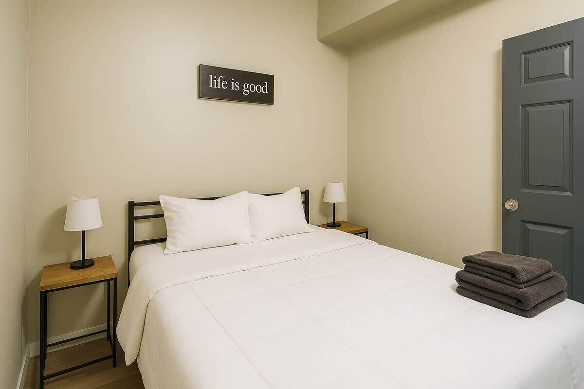 A neatly made bed with white bedding, two bedside lamps, a stack of folded towels, and a sign on the wall that reads 'life is good'.