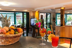 A table with a fruit basket, two drinks with fruit garnishes, and a bar area in the background. - Hotel Verdemare (Rimini)