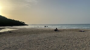 Una playa con gente caminando y nadando, cielo despejado y un paisaje montañoso al fondo.