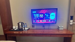 A wooden desk with a flat-screen TV displaying a streaming service interface, a kettle, two water bottles, and a chair. - Hotel Grand Matoa Timika (Timika)