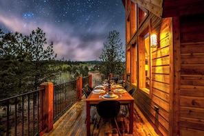 Eine Holzterrasse mit einem Esstisch, mit Blick auf eine bewaldete Landschaft unter einem Sternenhimmel.
