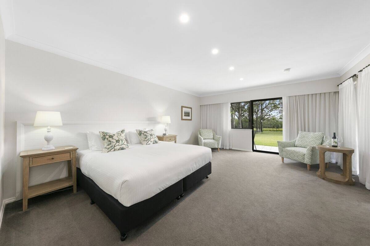 A spacious bedroom with a large bed, a nightstand, a chair, and a view of the outdoors through a large window.