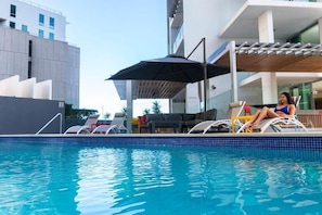 Ein Swimmingpool mit Liegestühlen und Sonnenschirm, umgeben von modernen Gebäuden.