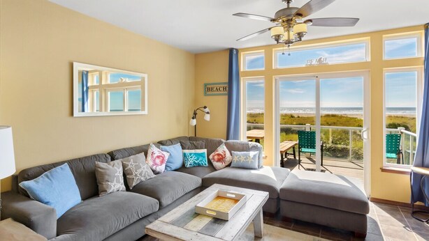A living room with a gray sectional sofa, a coffee table, a ceiling fan, and a view of the beach through large windows. - Beachfront Home w/ Hot Tub | Sleeps 9 | Dogs OK (Ocean Shores)