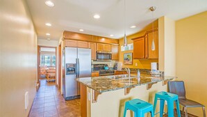 A kitchen with granite countertops, wooden cabinets, a stainless steel refrigerator, a microwave, and a sink. There are two blue bar stools and a chair. - Beachfront Home w/ Hot Tub | Sleeps 9 | Dogs OK (Ocean Shores)