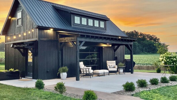 A dark wooden house with a covered patio, outdoor seating, and a view of a field.