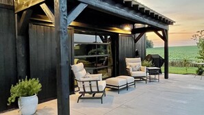 An outdoor patio with two white armchairs and a small table, a pergola structure, and a view of a green field.