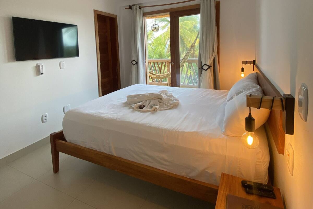 Un dormitorio con una cama, un televisor instalado en la pared, una mesita de noche con lámpara y una ventana con cortinas.