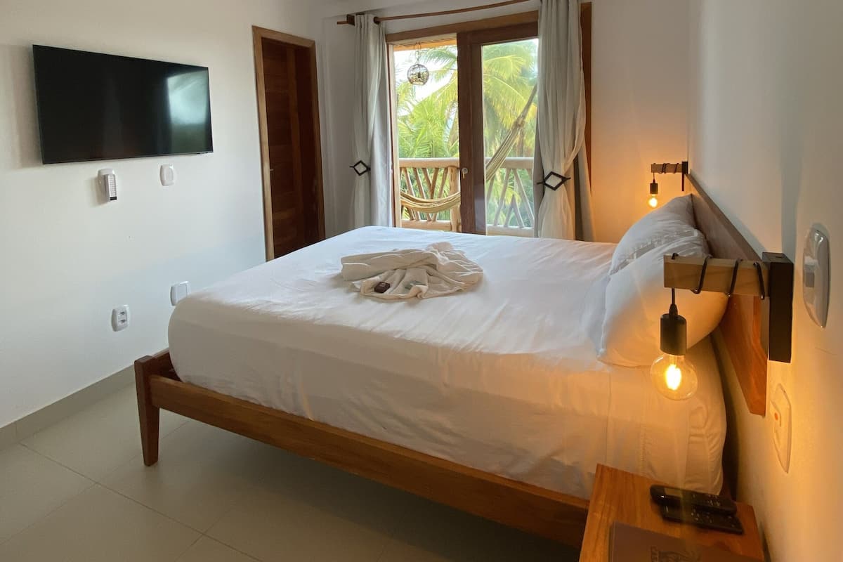 A bedroom with a bed, a TV mounted on the wall, a bedside table with a lamp, and a window with curtains.
