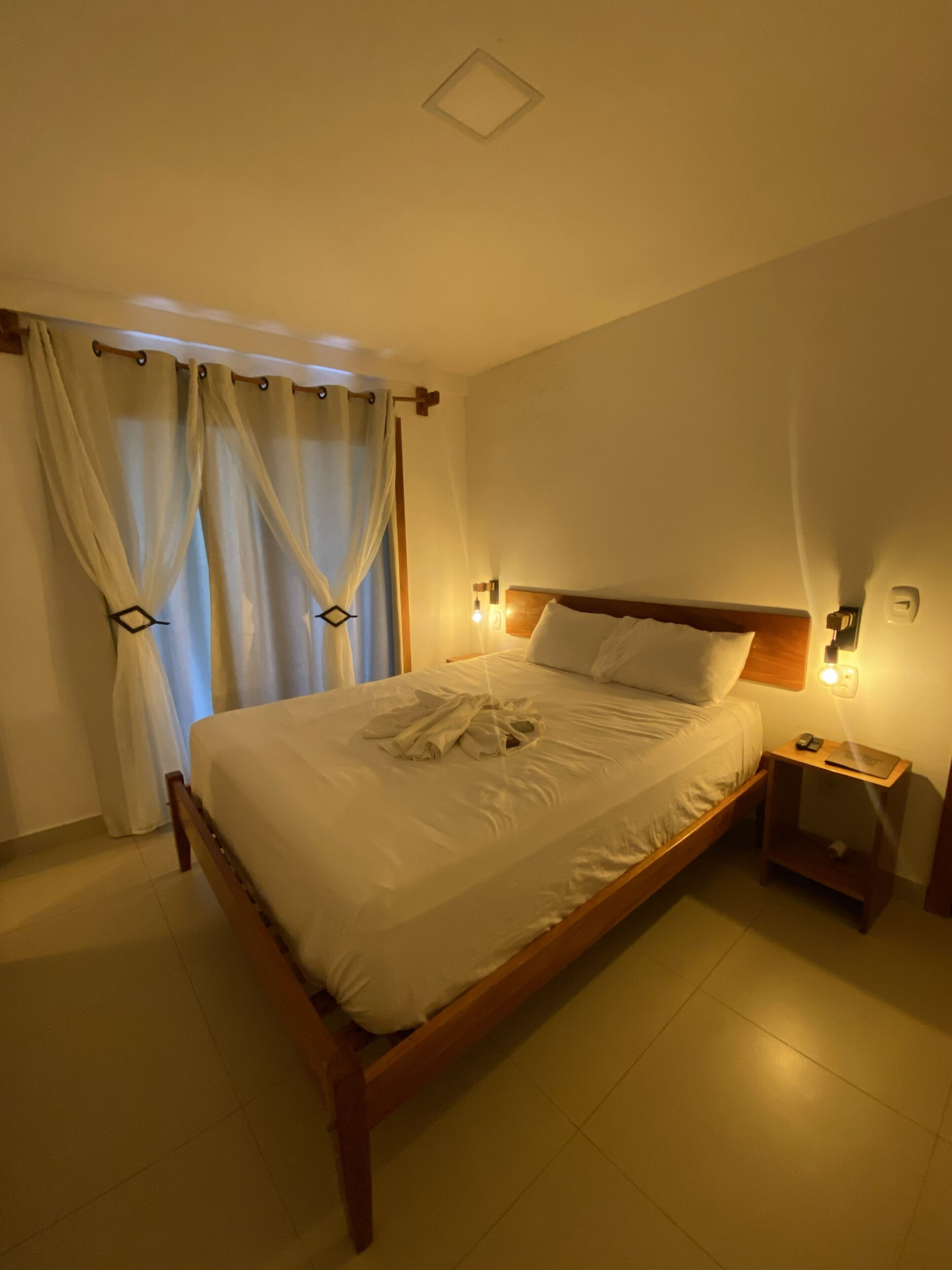 Un letto rifatto con un'ossatura in legno, un comodino con una lampada e una finestra con tende.