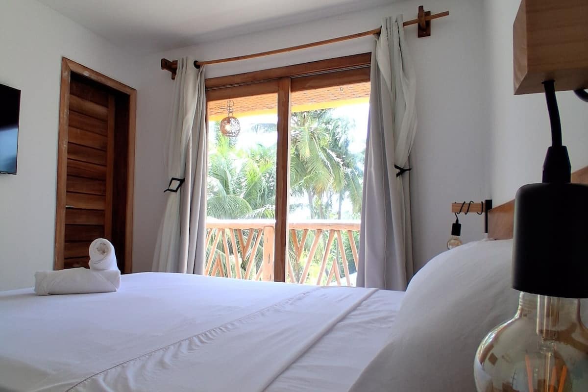 Habitación de hotel con cama, puerta de madera, ventana con cortinas y vista a palmeras.