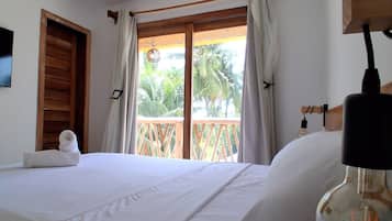 Habitación de hotel con cama, puerta de madera, ventana con cortinas y vista a palmeras.