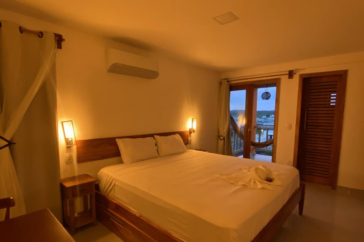 Un dormitorio con una cama grande, mesitas de noche, una silla de madera, una ventana con cortinas y vistas al exterior.
