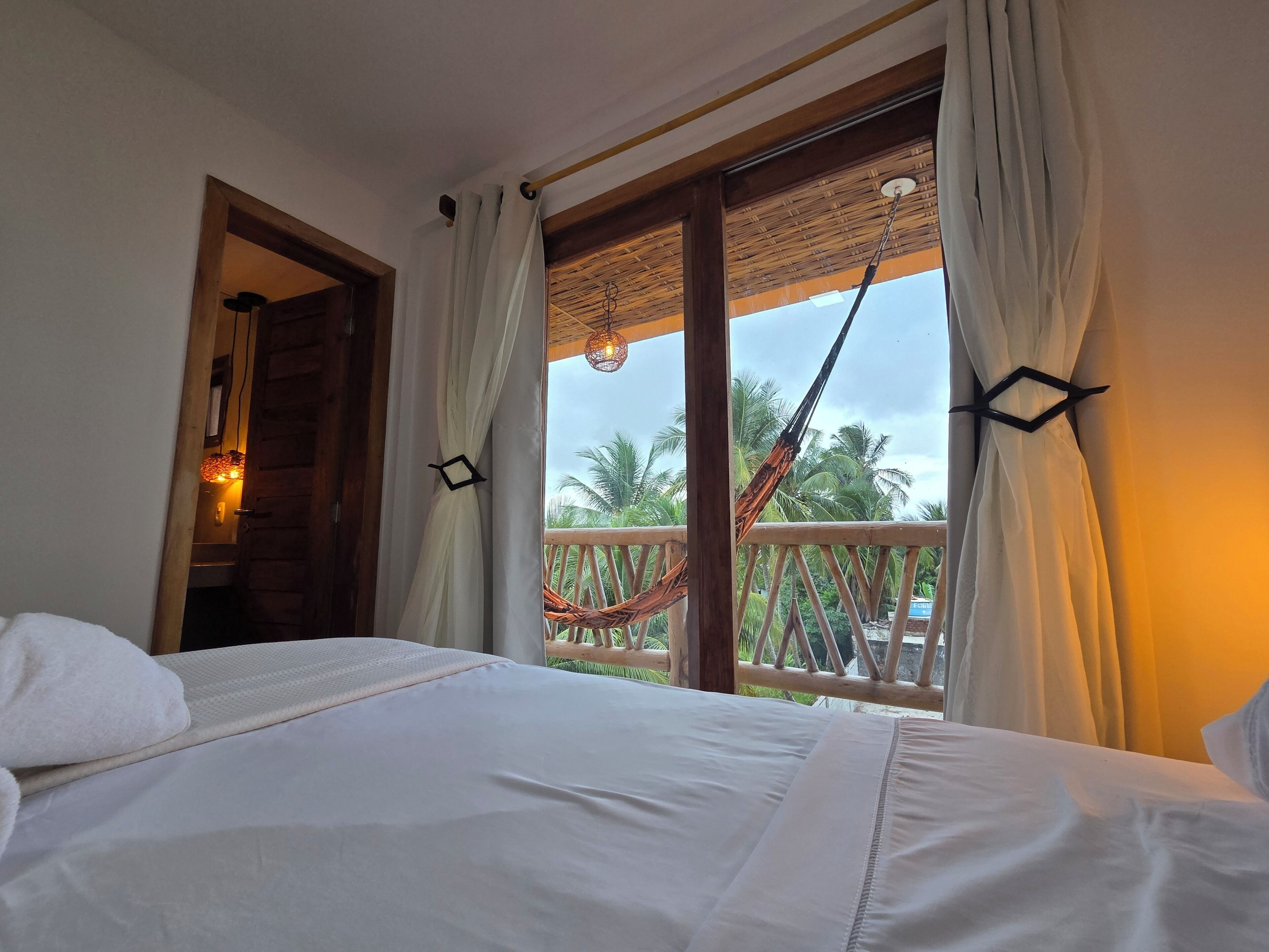 A hotel room with a bed, curtains, a view of palm trees, and a hammock.