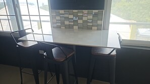 Une cuisine moderne avec un plan de travail blanc, deux tabourets de bar et une vue sur l’extérieur grâce à de grandes fenêtres.