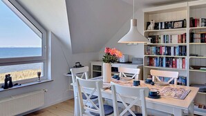 Ein Zimmer mit großem Fenster, einem Esstisch, Stühlen, einem Bücherregal und einer Deckenleuchte.